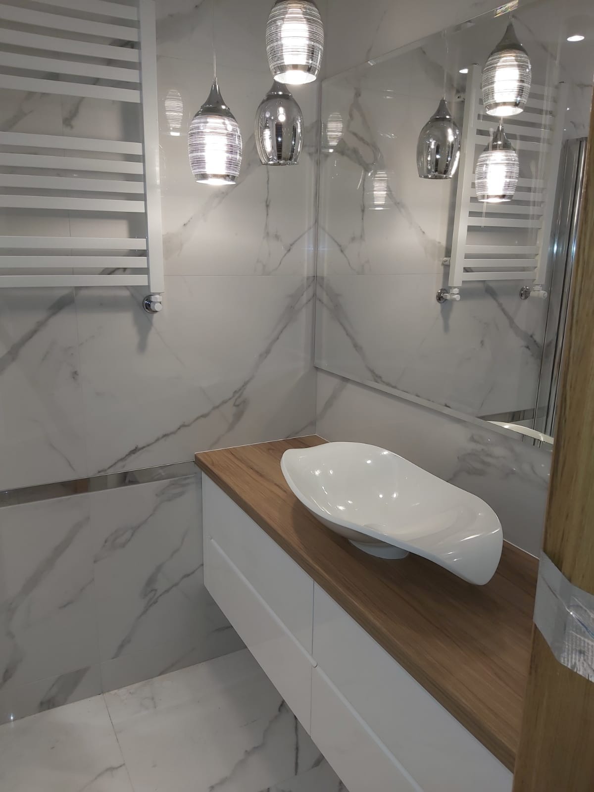 Nowoczesna łazienka z marmurowymi płytkami, wiszącą szafką z drewnianym blatem i asymetryczną umywalką oraz designerskimi lampami wiszącymi.