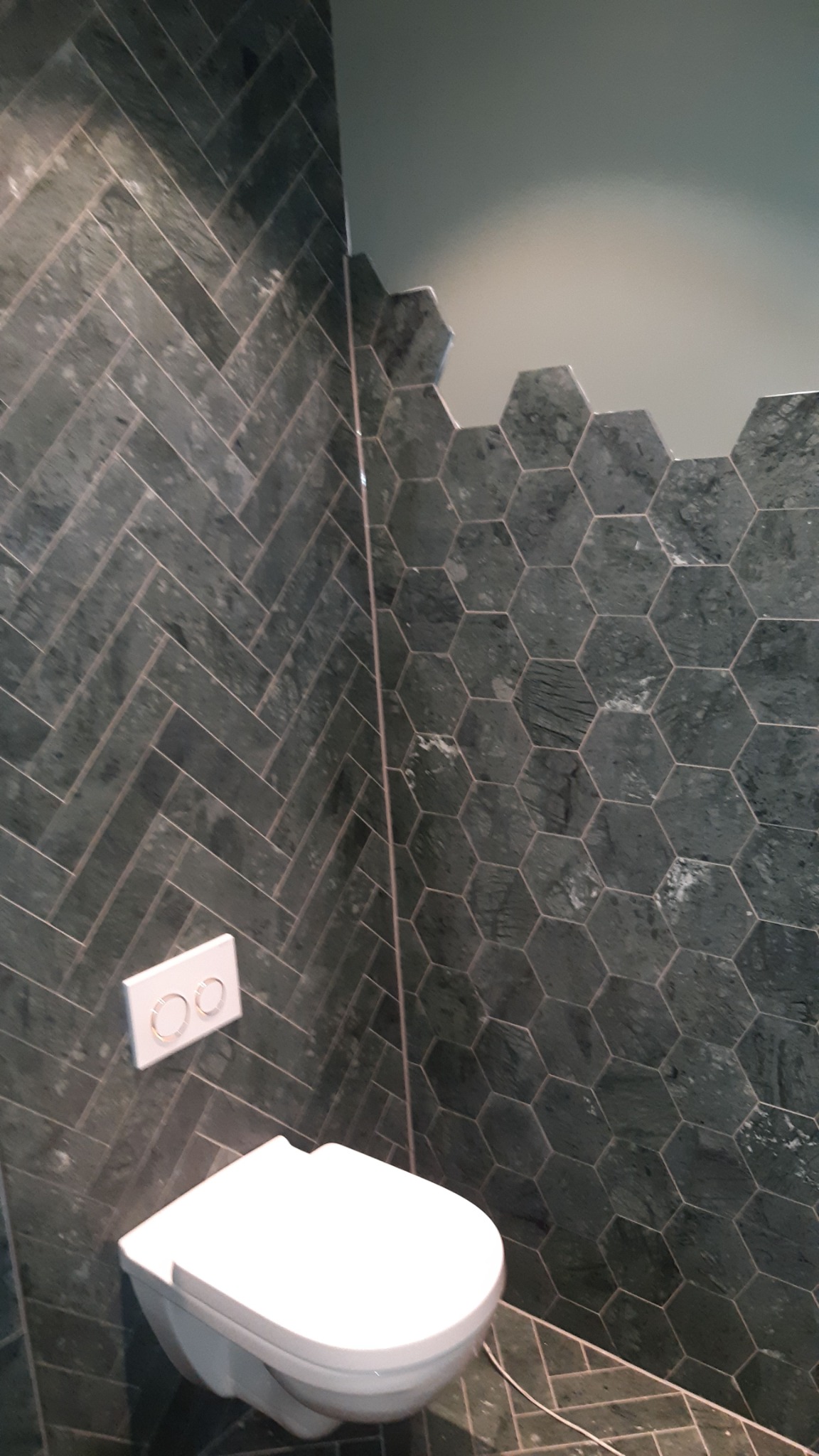 Nowoczesna toaleta podwieszana z przyciskiem spłukującym na ścianie wyłożonej ciemnozielonymi płytkami w jodełkę i heksagony.