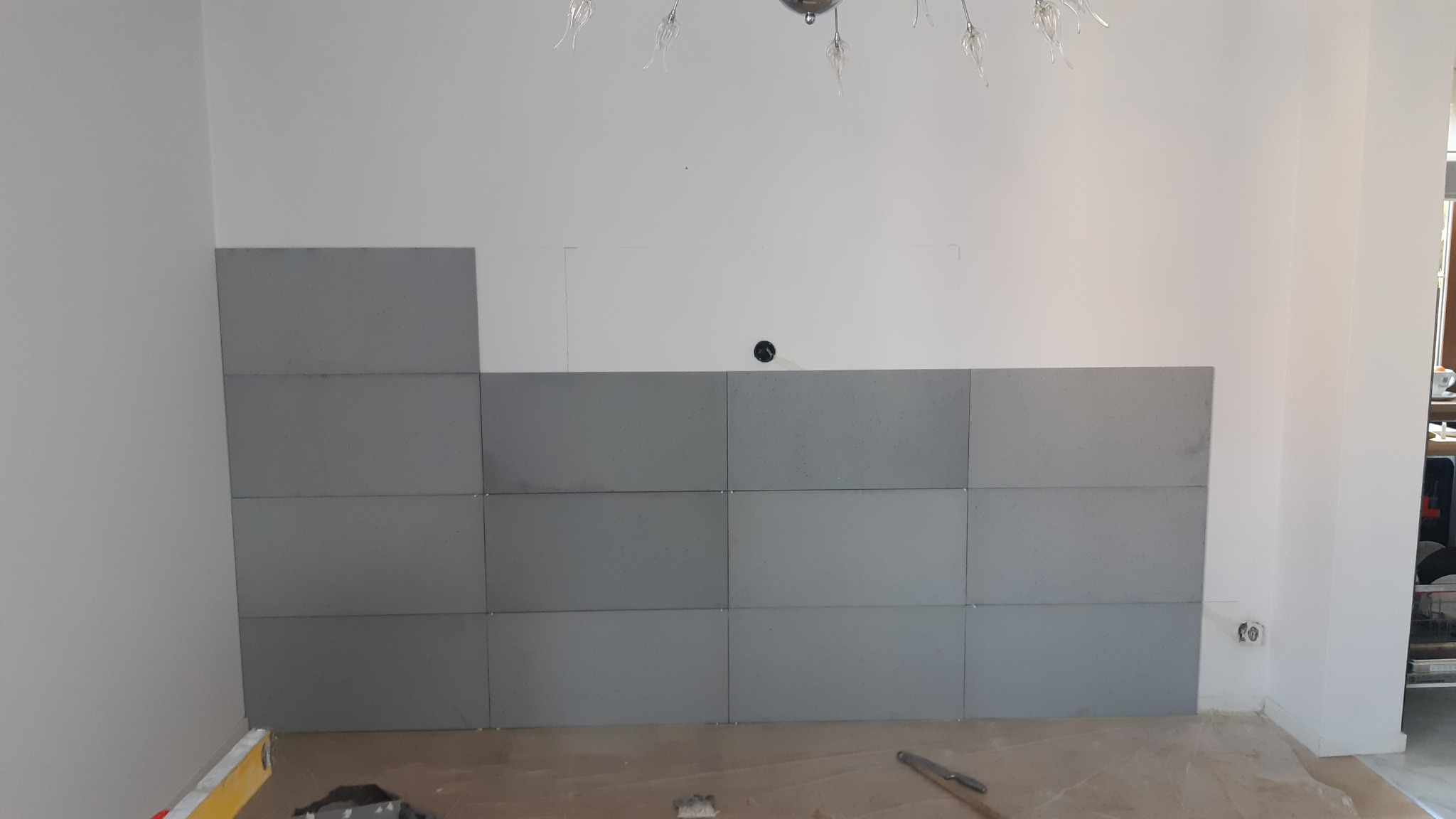 Montaż szarych płytek ściennych w trakcie prac wykończeniowych, widoczny poziomica i narzędzia na podłodze zabezpieczonej folią, brakująca płytka w górnym rogu.