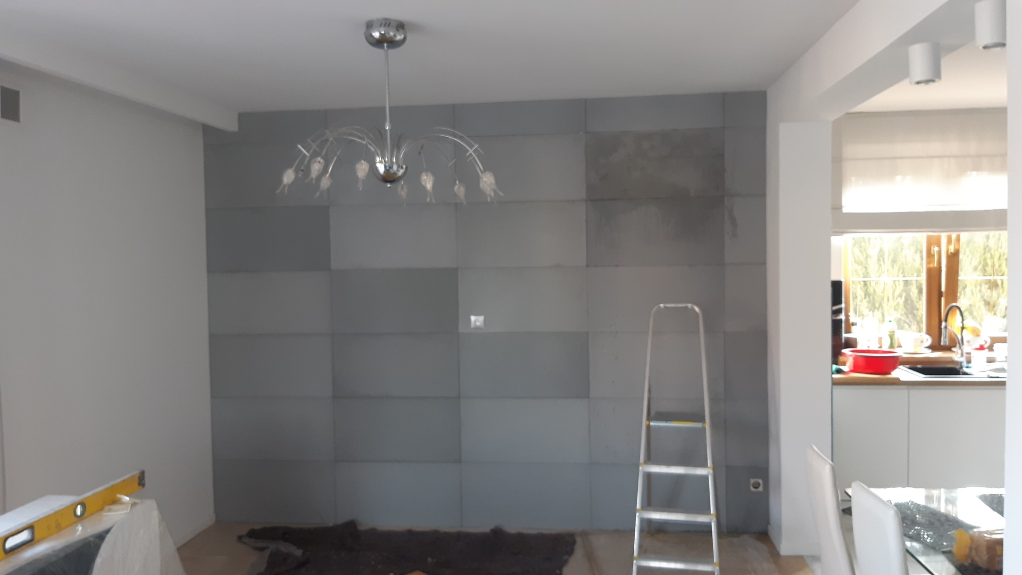 Ściana wykończona szarymi panelami, widoczna drabina i poziomica, w tle kuchnia.