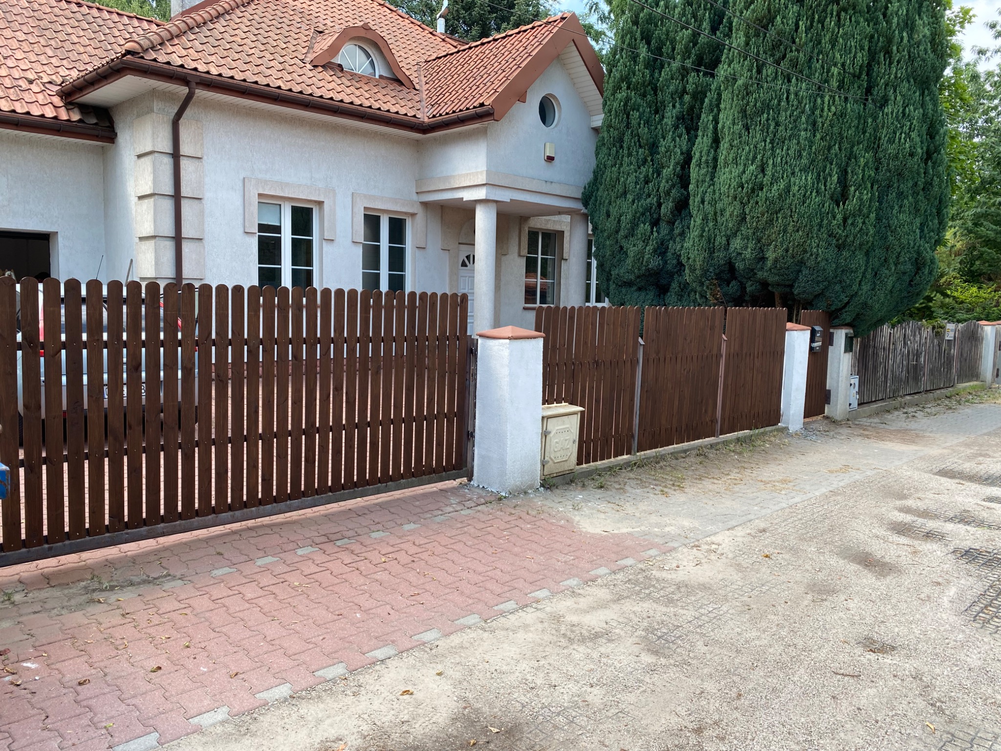 Dom jednorodzinny z brązowym drewnianym ogrodzeniem panelowym i bramą przesuwną, widoczne kostki brukowe na podjeździe oraz skrzynka gazowa przy słupku ogrodzenia.
