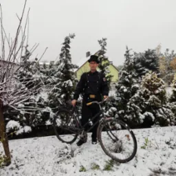 Mężczyzna w tradycyjnym stroju, z cylindrem i szczotkami, stoi obok roweru na zaśnieżonym podwórku, na tle iglaków i domu.