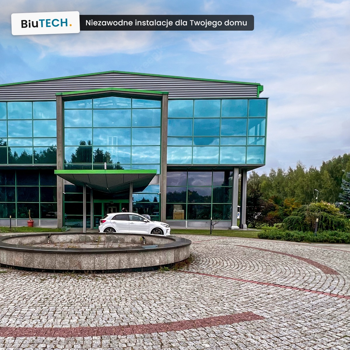 Nowoczesny budynek biurowy z przeszkloną fasadą i logo BiuTECH, na pierwszym planie fontanna i brukowany plac.