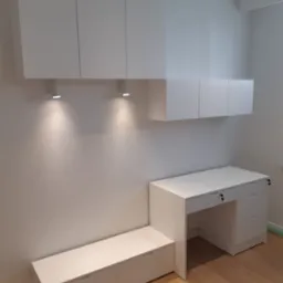 Białe meble biurowe na wymiar: wiszące szafki, biurko z szufladami zamykanymi na klucz i niska szafka z szufladami, oświetlenie punktowe, parkiet na podłodze.