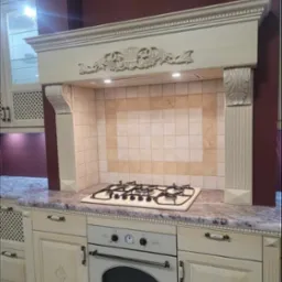 Klasyczna kuchnia w odcieniach beżu i bordo, z frezowanymi frontami szafek, marmurowym blatem i ozdobnym okapem nad płytą gazową.