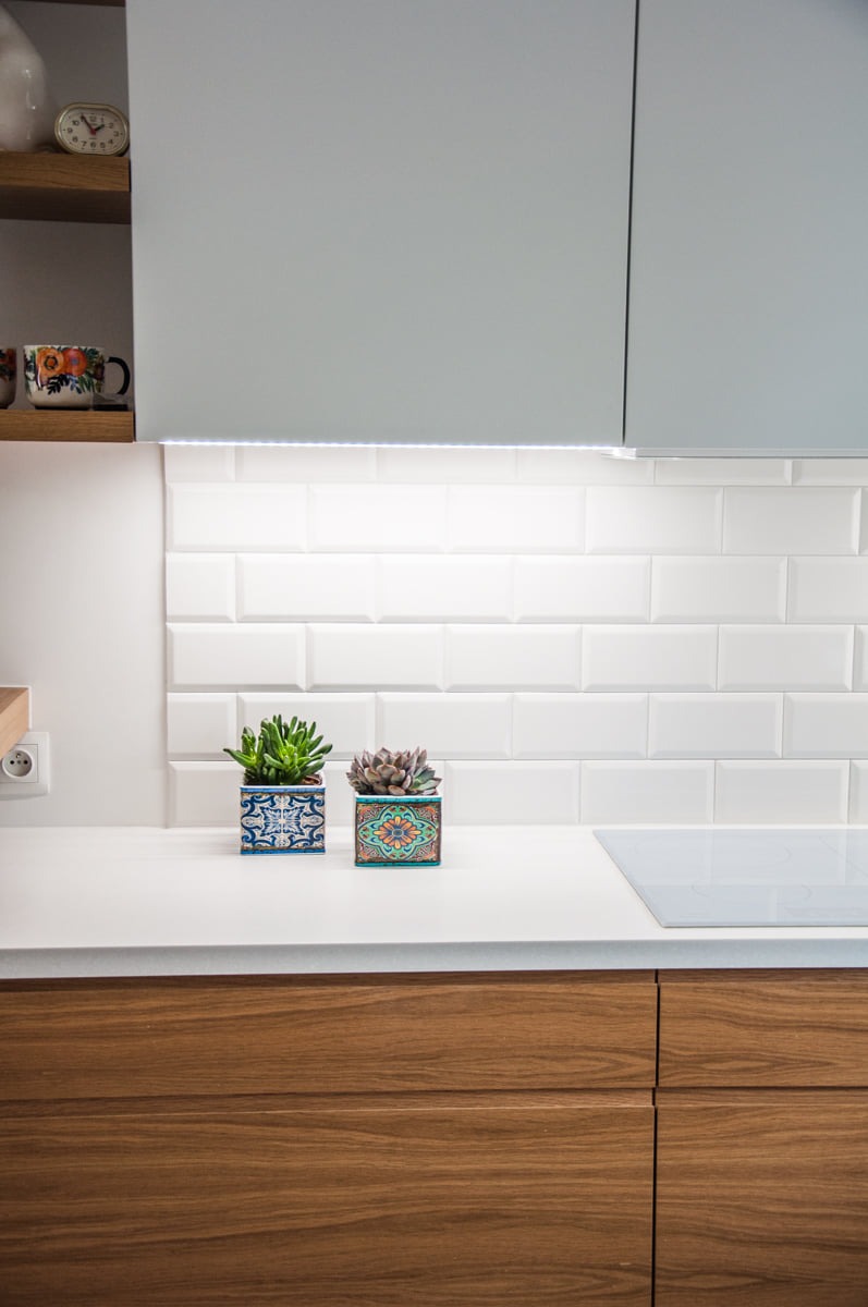Nowoczesna kuchnia: białe płytki typu metro, drewniane fronty szafek, biały blat, dwie małe sukulenty w ozdobnych doniczkach, oświetlenie podszafkowe LED.