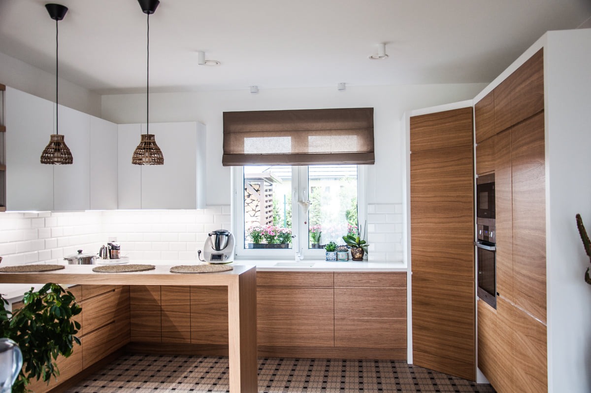 Nowoczesna kuchnia w Gdyni z drewnianymi blatami i frontami, białymi szafkami wiszącymi i wzorzystą podłogą. W oknie widoczne kwiaty w doniczkach, a nad blatem wiszą plecione lampy.