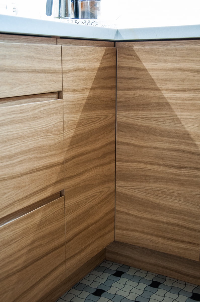 Fragment narożnej zabudowy kuchennej z jasnego drewna z szufladami i blatem z białego konglomeratu, widoczna mozaika podłogowa.