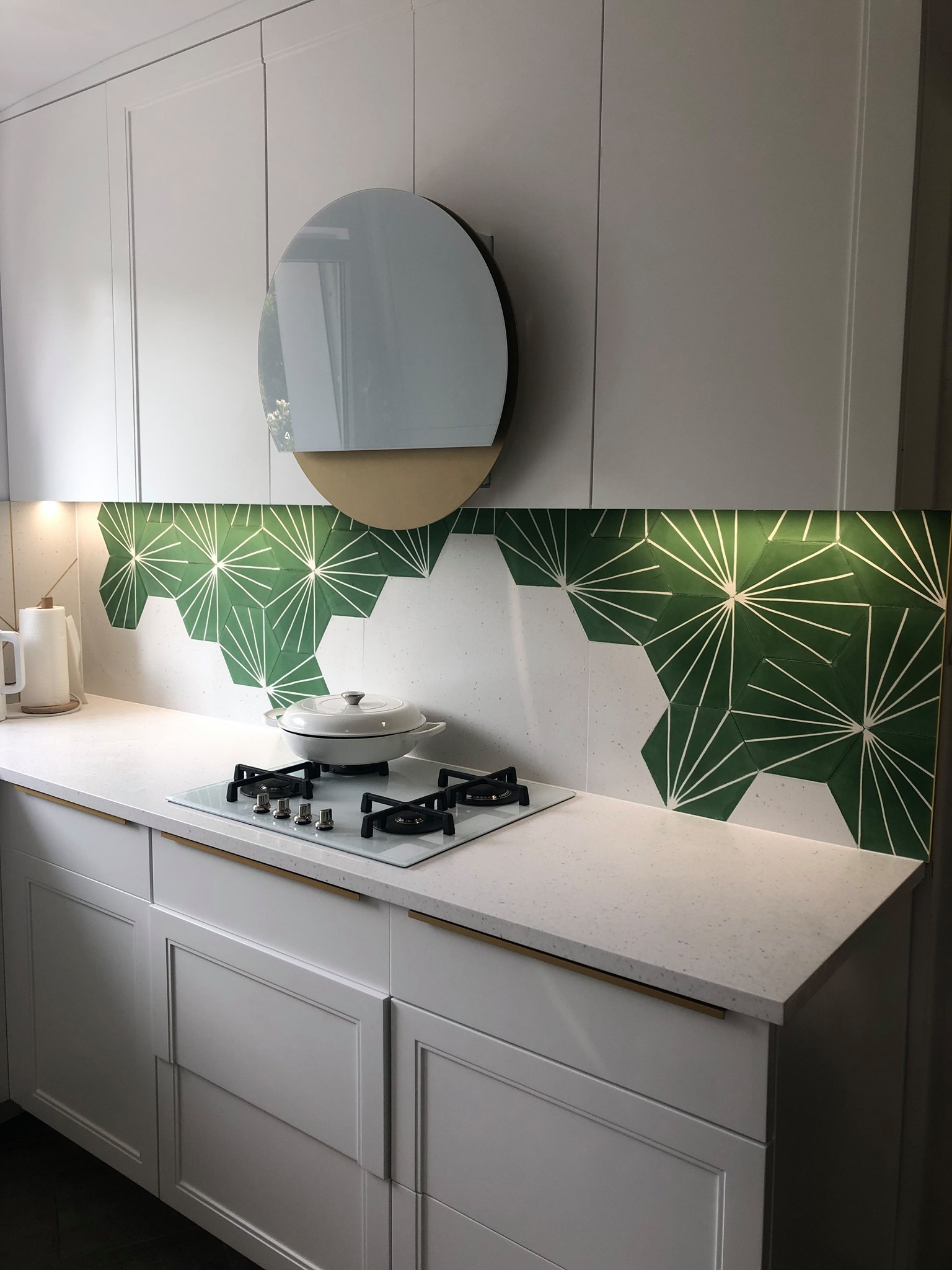 Biała kuchnia z zielonymi heksagonalnymi płytkami nad blatem, okrągły wyciąg, biała płyta grzewcza z garnkiem, minimalistyczne szafki z podłużnymi złotymi uchwytami.