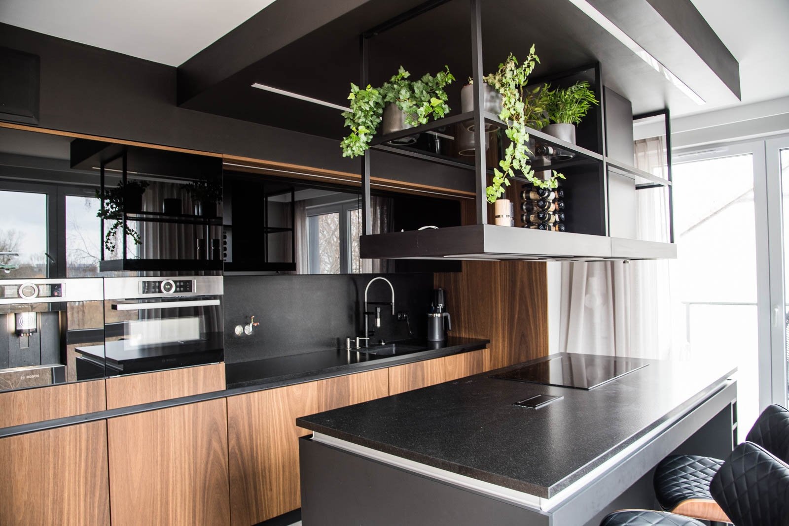 Nowoczesna kuchnia z drewnianymi frontami, czarnym blatem i wiszącą półką z roślinami doniczkowymi i butelkami wina.
