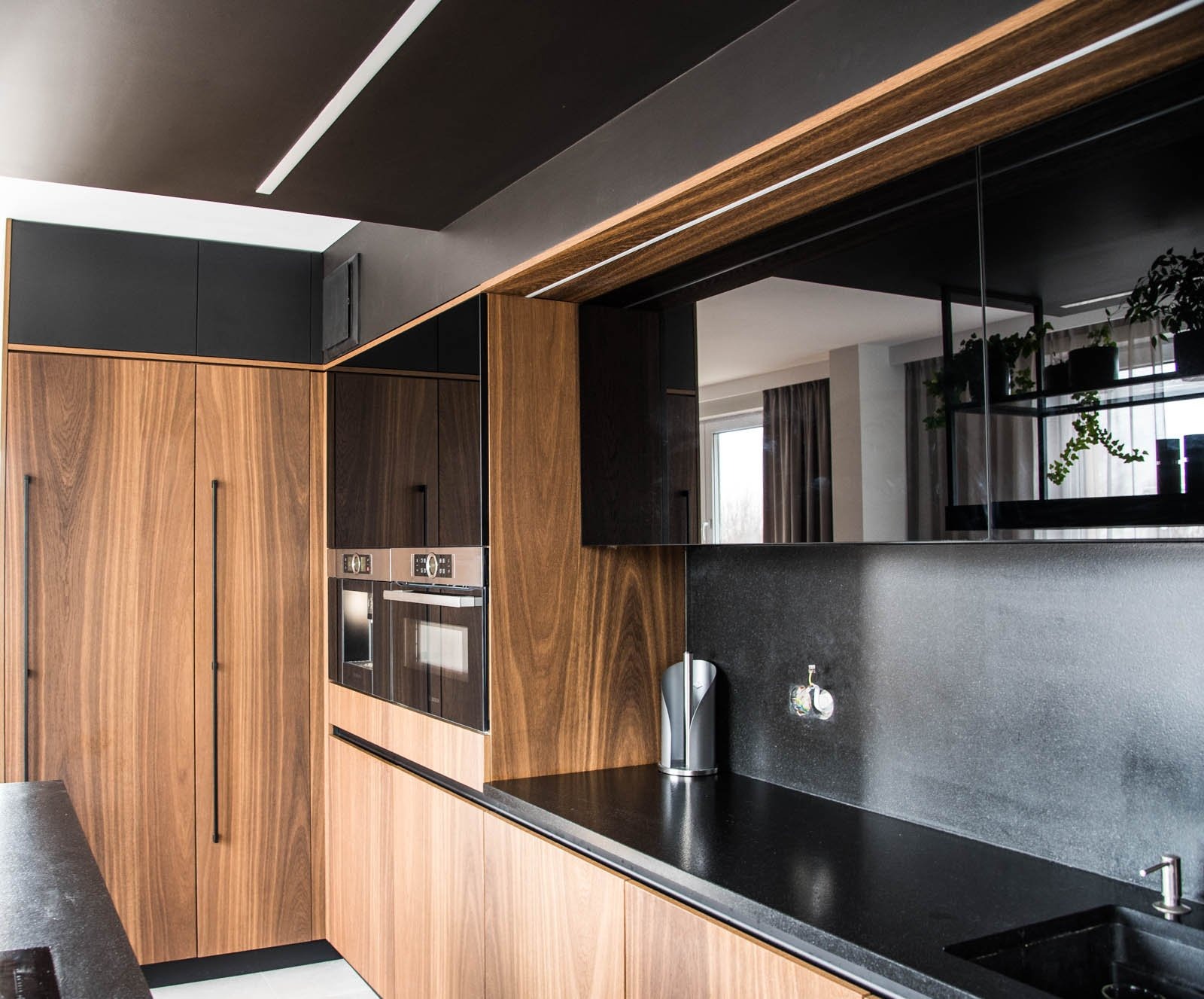 Nowoczesna kuchnia z drewnianymi frontami szafek, czarnym blatem i zabudową pod sufit, z widocznymi piekarnikami i elementami dekoracyjnymi w odbiciu.