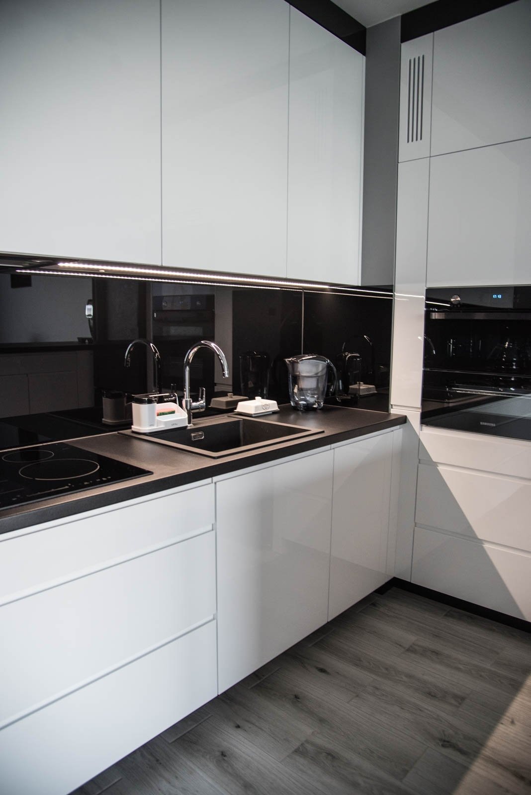 Nowoczesna kuchnia z białymi, lakierowanymi frontami, czarnym blatem i szklanym panelem nad blatem z oświetleniem LED, widoczny zlew, bateria i płyta indukcyjna.