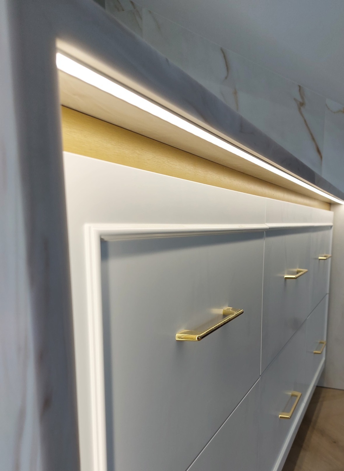 Szare szafki z frezowanymi frontami i złotymi uchwytami, oświetlone listwą LED, widok z dołu, fragment marmurowej ściany w tle.