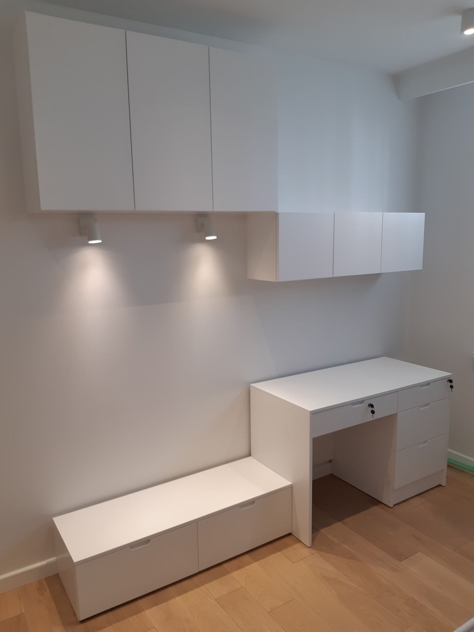 Białe meble biurowe na wymiar: wiszące szafki, biurko z szufladami zamykanymi na klucz i niska szafka z szufladami, oświetlenie punktowe, parkiet na podłodze.