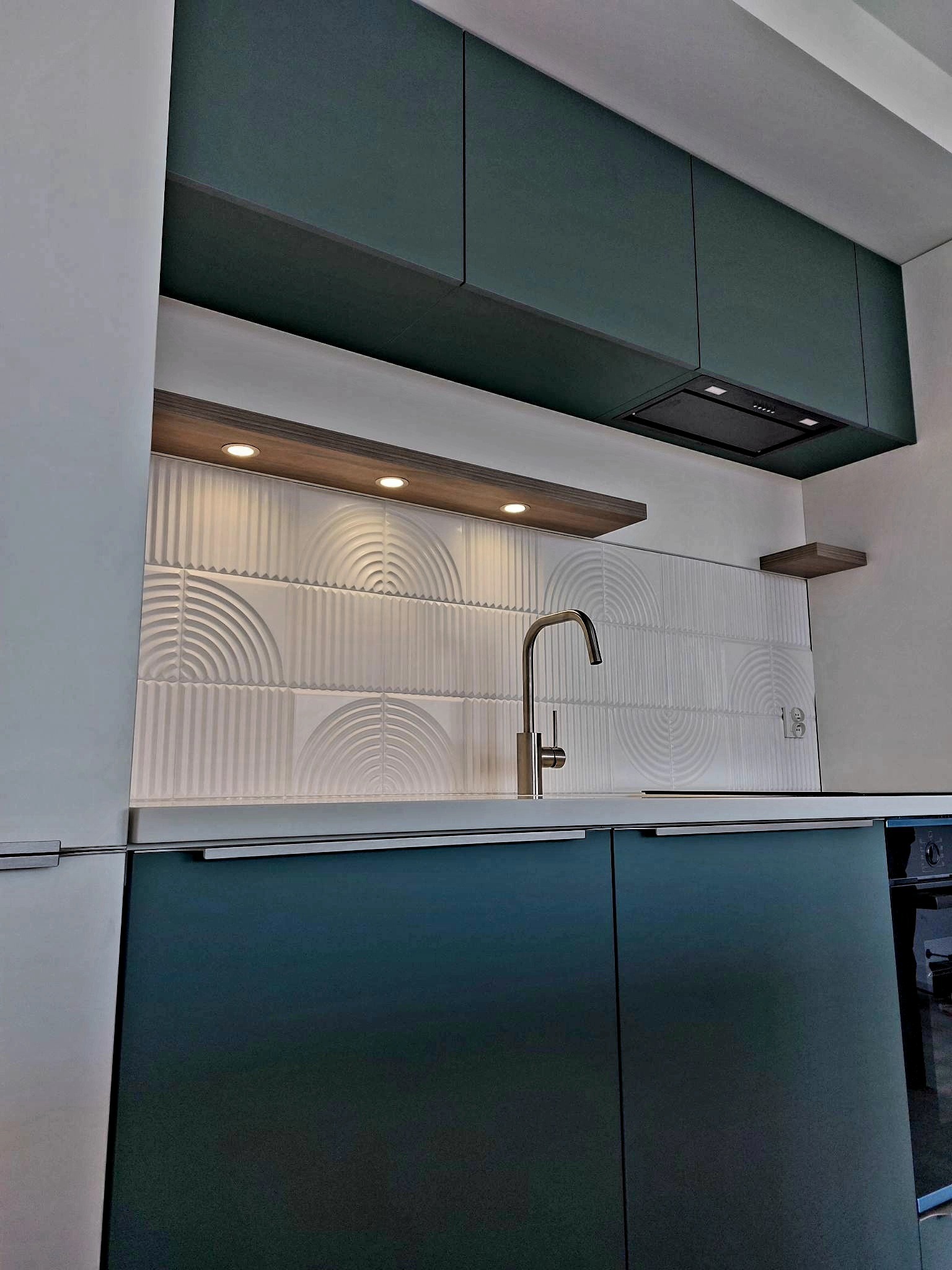 Nowoczesna kuchnia z ciemnozielonymi szafkami, białymi płytkami dekoracyjnymi, drewnianą półką z oświetleniem punktowym i chromowaną baterią.