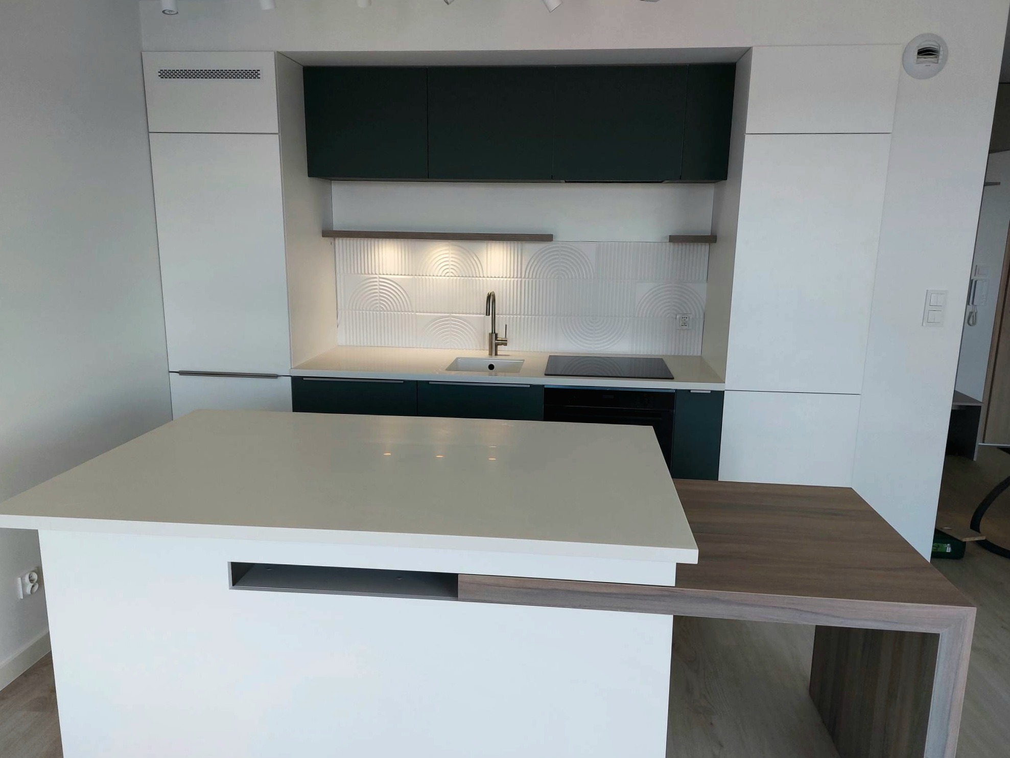 Nowoczesna kuchnia z biało-zielonymi szafkami, wyspą z białym blatem i drewnianym przedłużeniem, oraz dekoracyjnymi białymi płytkami na ścianie.