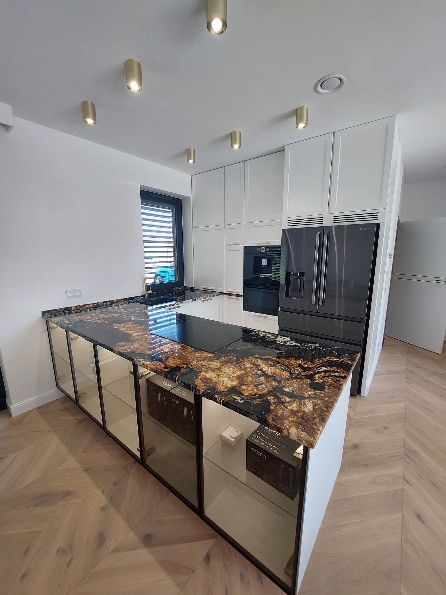 Nowoczesna kuchnia z wyspą, blat z ciemnego kamienia z brązowymi żyłami, białe szafki z frontami lustrzanymi, parkiet w jodełkę, oświetlenie punktowe, czarna lodówka Samsung.