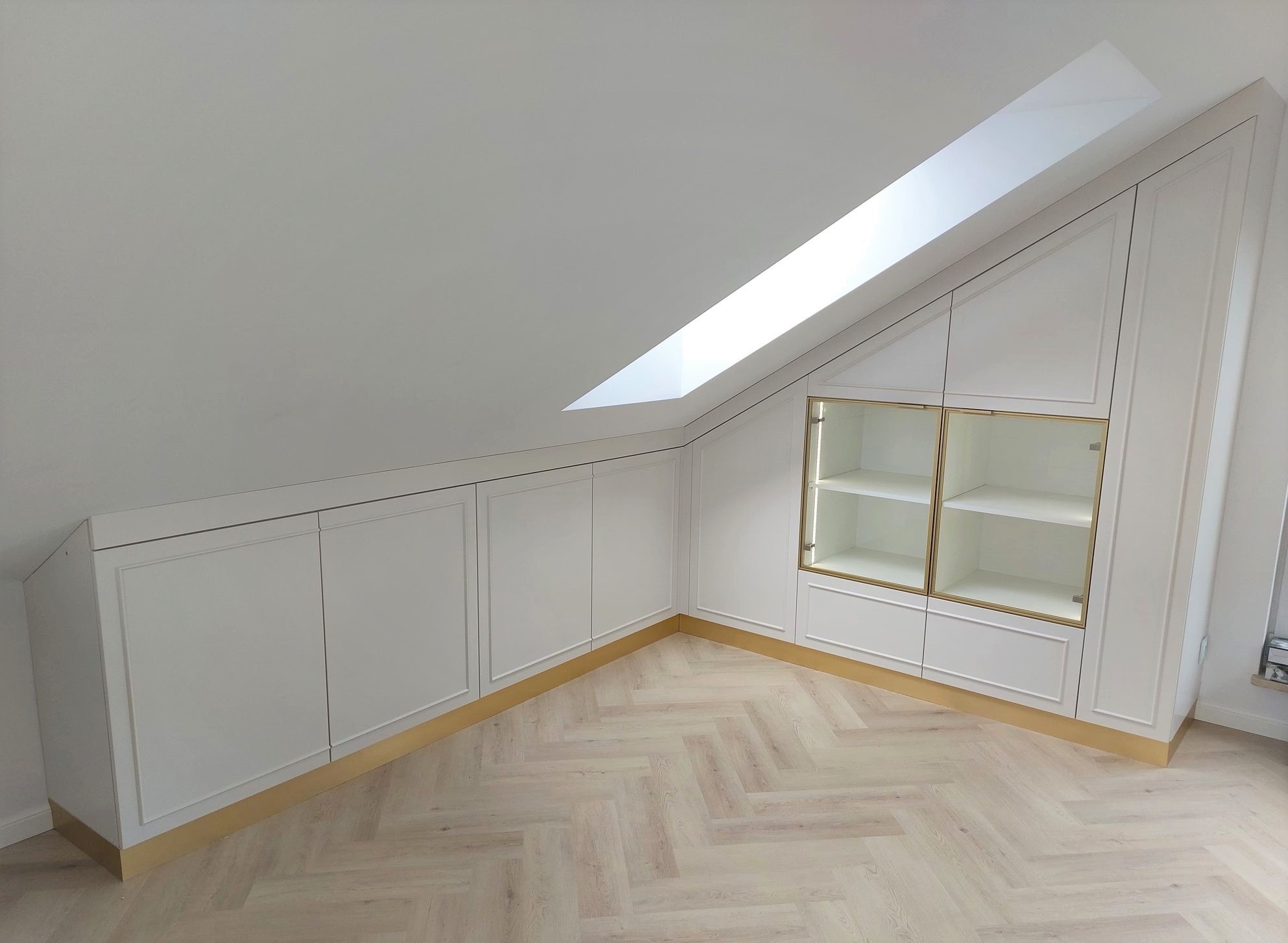 Biała zabudowa meblowa pod skosem z przeszkloną witryną, złote akcenty, parkiet w jodełkę, oświetlenie wewnątrz witryny