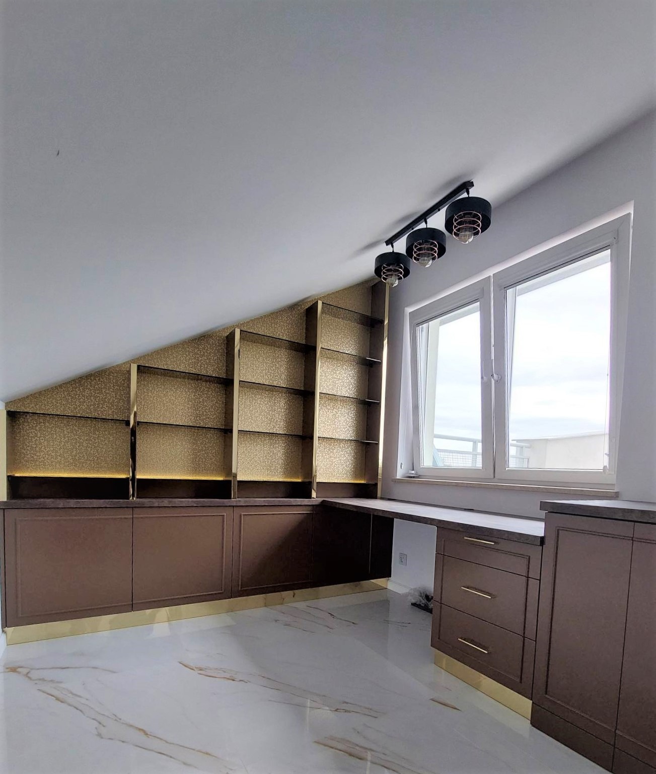 Zabudowa wnęki na poddaszu z brązowymi szafkami, złotymi akcentami i oświetlonymi półkami, uzupełniona blatem roboczym i trzema lampami w stylu industrialnym.