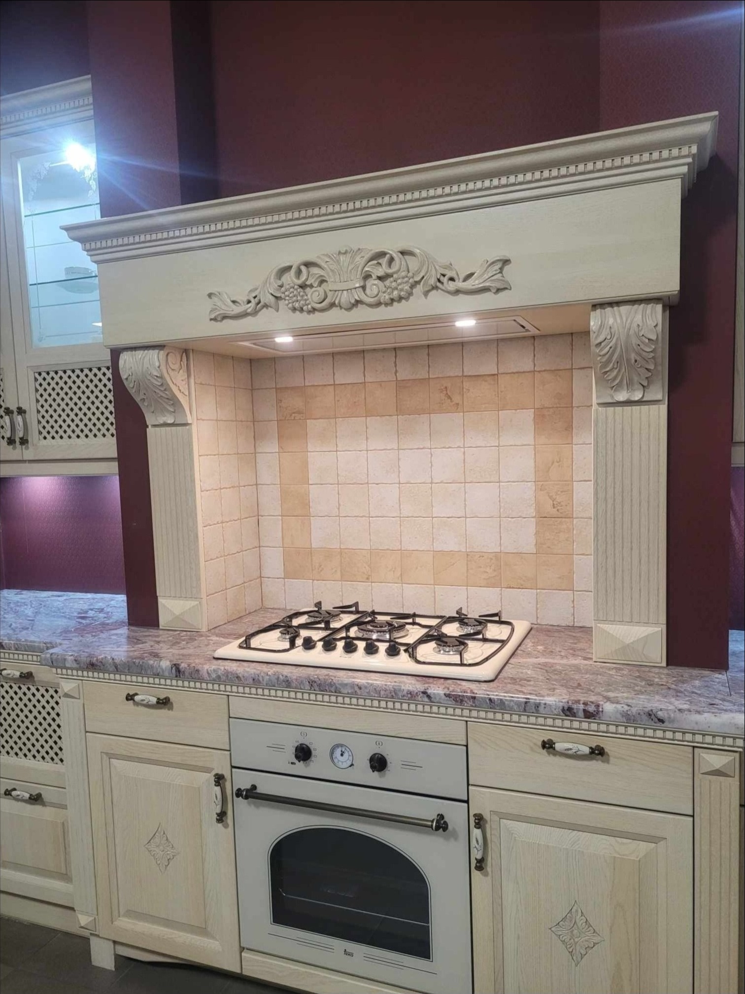 Klasyczna kuchnia w odcieniach beżu i bordo, z frezowanymi frontami szafek, marmurowym blatem i ozdobnym okapem nad płytą gazową.