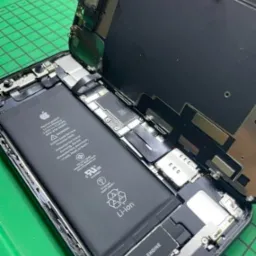 Rozłożony iPhone z widoczną baterią, płytą główną i elementami mocującymi ekran, leżący na zielonej macie warsztatowej.