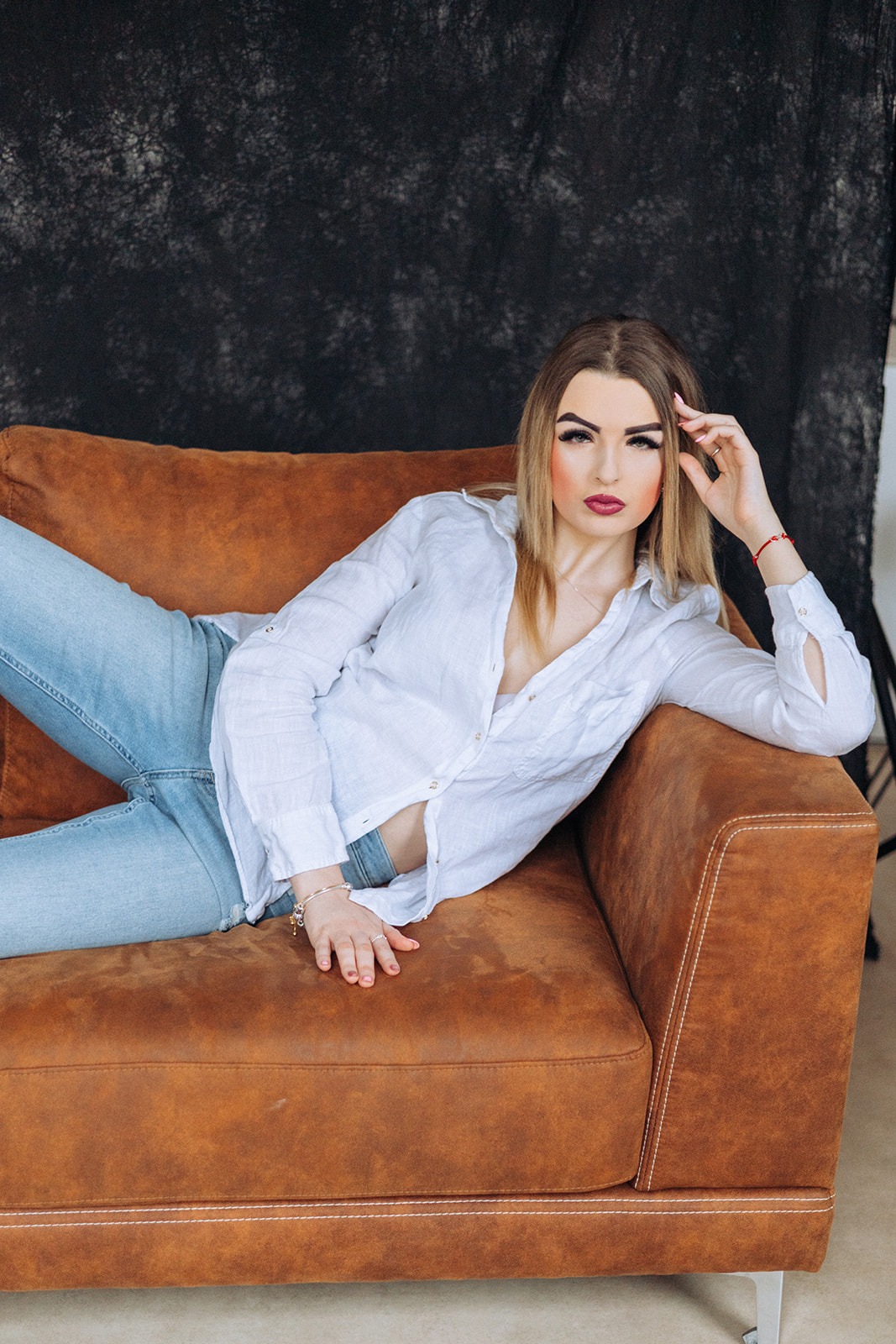Portret kobiety w białej koszuli i jeansach, leżącej na brązowej sofie na tle ciemnego tła.