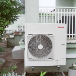 Biała zewnętrzna jednostka pompy ciepła Toshiba zamontowana na ścianie domu, widoczny wentylator, rurki odprowadzające skropliny i ozdobny płotek w tle.