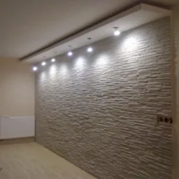 Ściana z dekoracyjnej cegły w salonie, oświetlona punktowymi lampami sufitowymi. Widoczne gniazdka elektryczne. Podłoga wyłożona panelami.