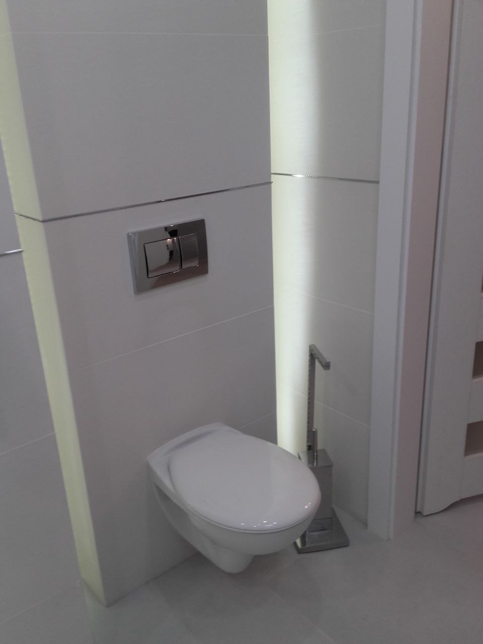 Nowoczesna toaleta podwieszana z chromowanym przyciskiem spłukiwania, w minimalistycznej łazience w Krakowie. Białe płytki i oświetlenie LED.