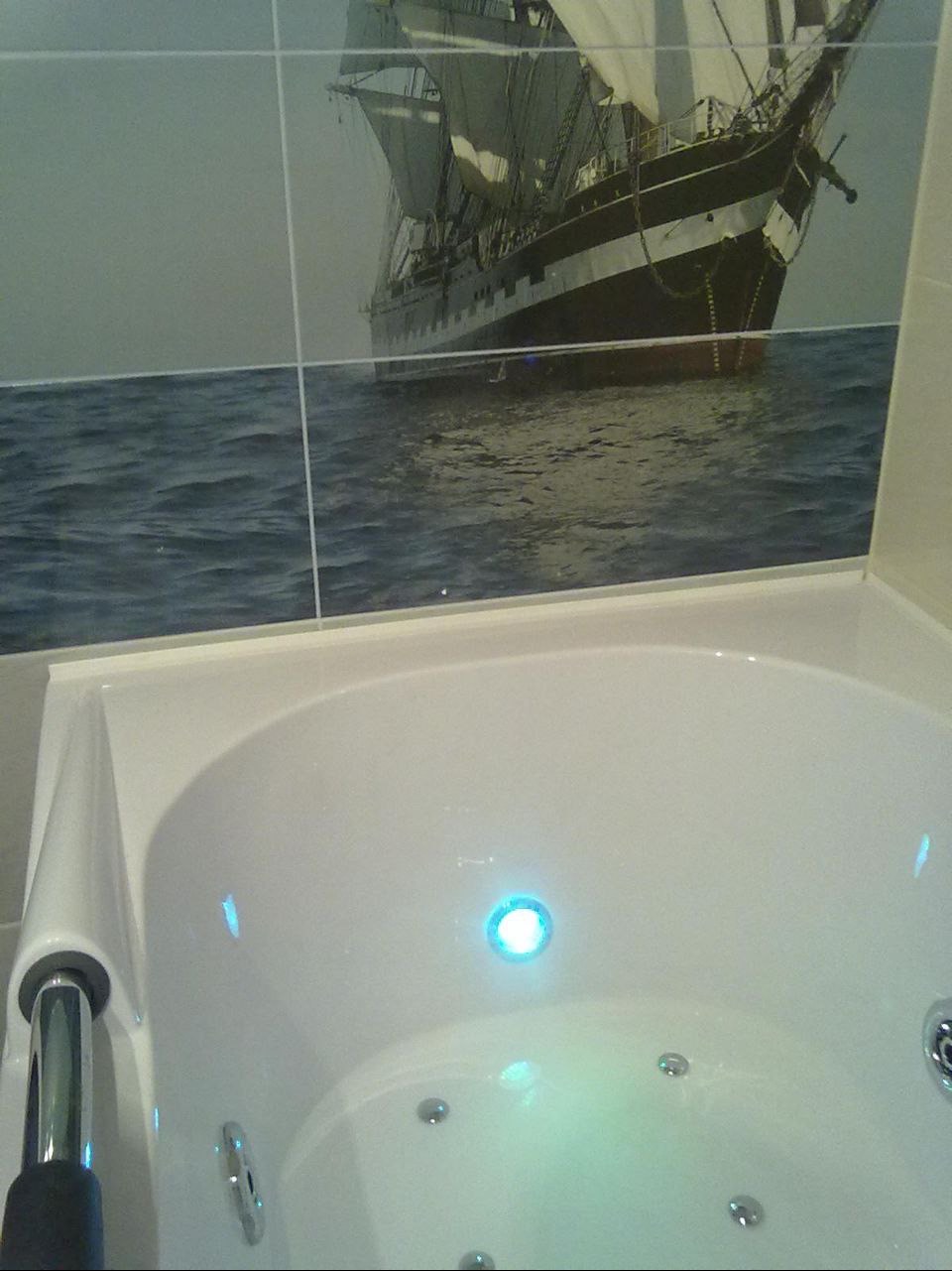 Biała wanna z hydromasażem i podświetleniem LED, na ścianie płytki z motywem żaglowca. Wykończenie łazienki w stylu morskim.