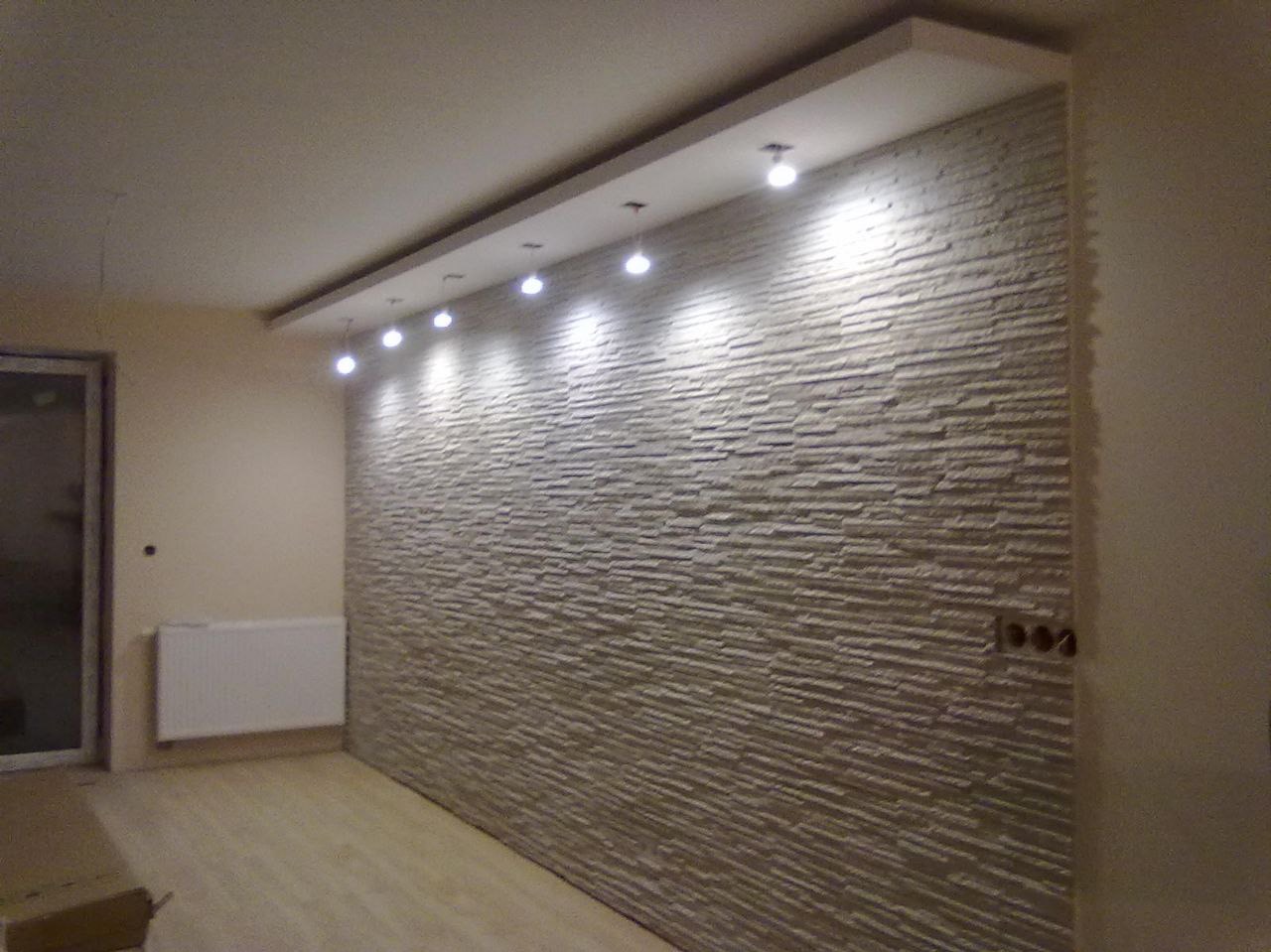 Ściana z dekoracyjnej cegły w salonie, oświetlona punktowymi lampami sufitowymi. Widoczne gniazdka elektryczne. Podłoga wyłożona panelami.