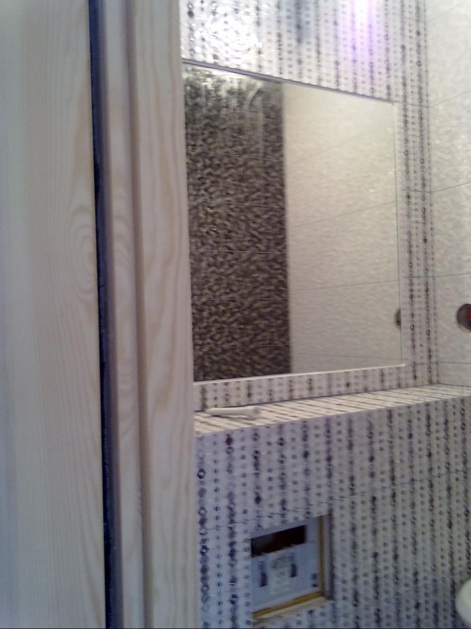 Wykończona łazienka z lustrem, wzorzystymi płytkami i drewnianym elementem. Widoczny fragment ściany z ciemniejszymi płytkami odbity w lustrze.