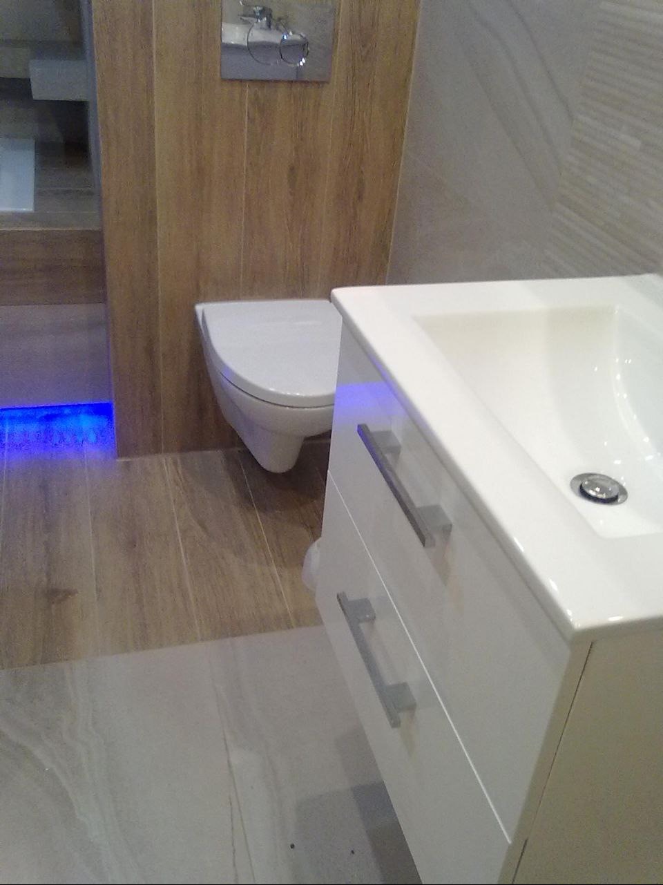 Nowoczesna łazienka z podświetleniem LED: biała umywalka z szafką, podwieszana toaleta i płytki imitujące drewno, tworzące spójną aranżację.