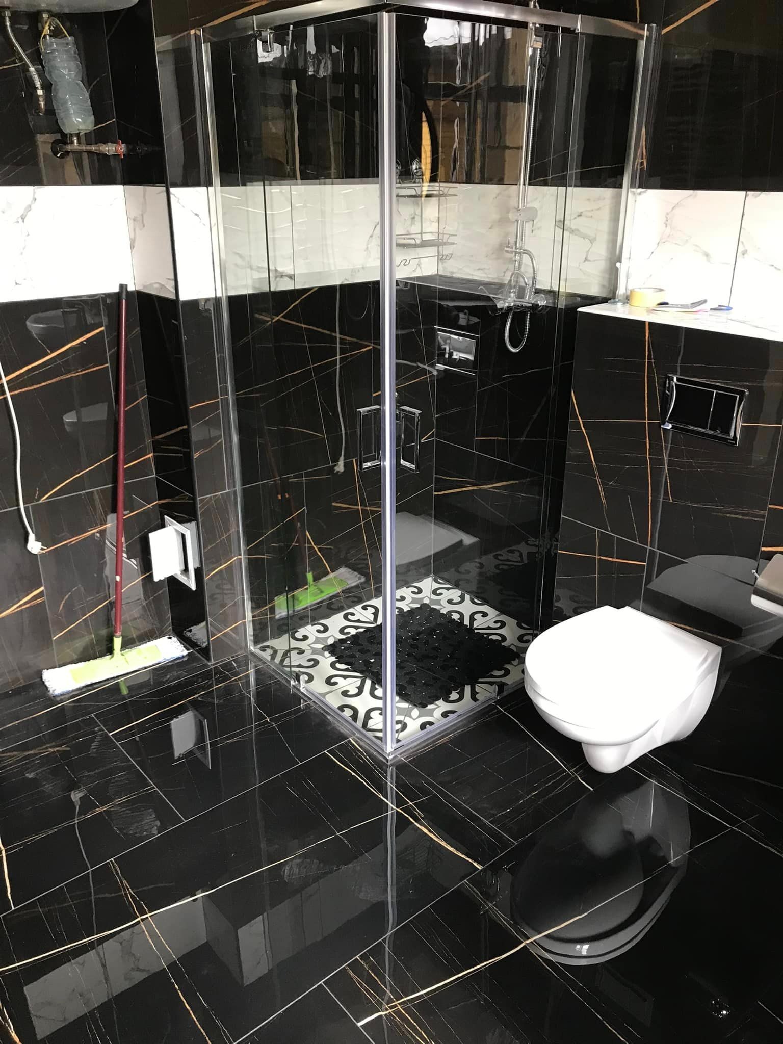 Wykończona łazienka w Gromadce z czarnymi płytkami imitującymi marmur ze złotymi żyłami, kabina prysznicowa ze szklanymi ściankami, biała toaleta i mop oparty o ścianę.