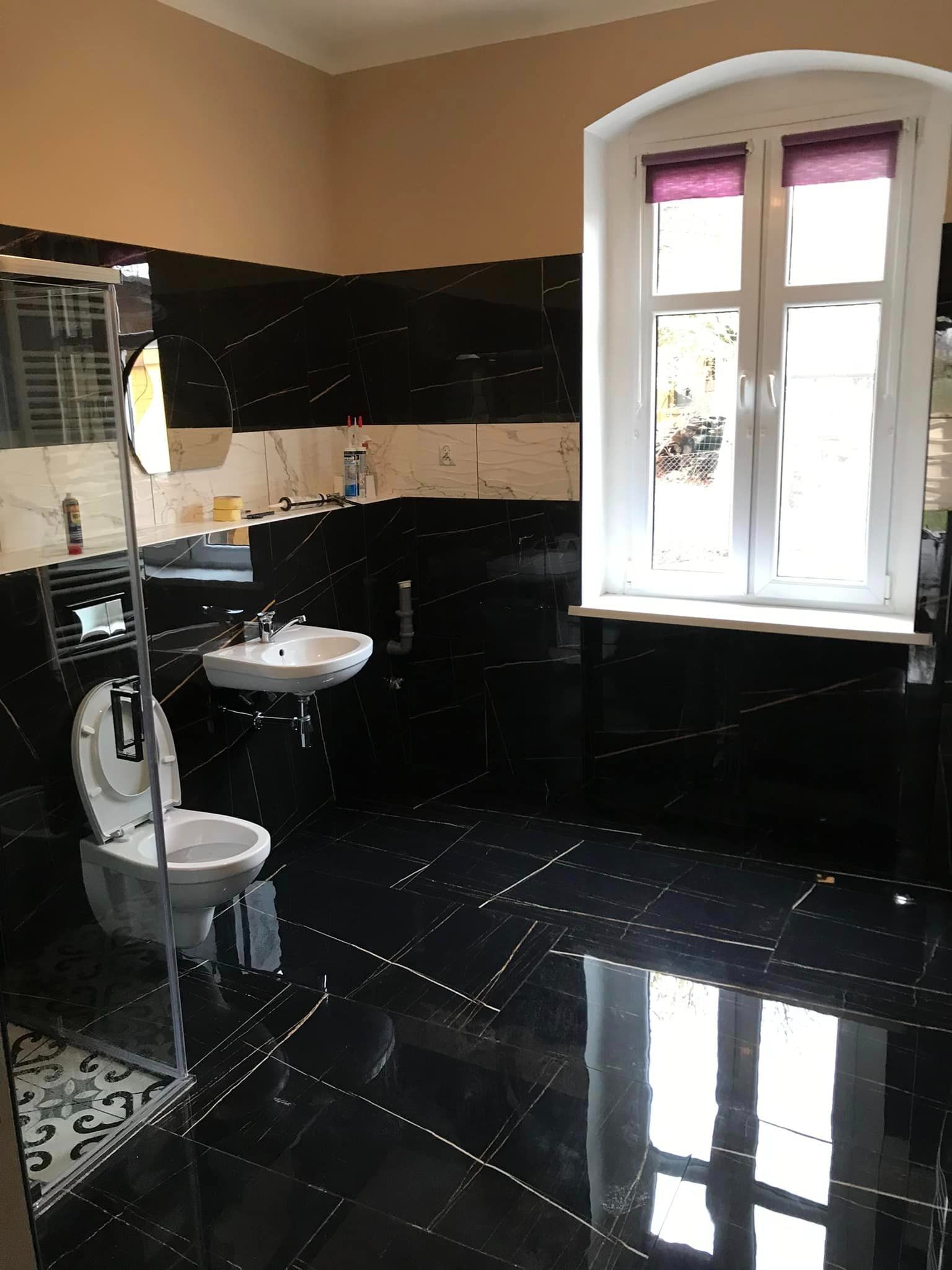 Wykończona łazienka z czarnymi płytkami imitującymi marmur, białą ceramiką sanitarną i oknem z roletami w kolorze fioletowym. Na półce nad umywalką widoczne akcesoria i narzędzia.