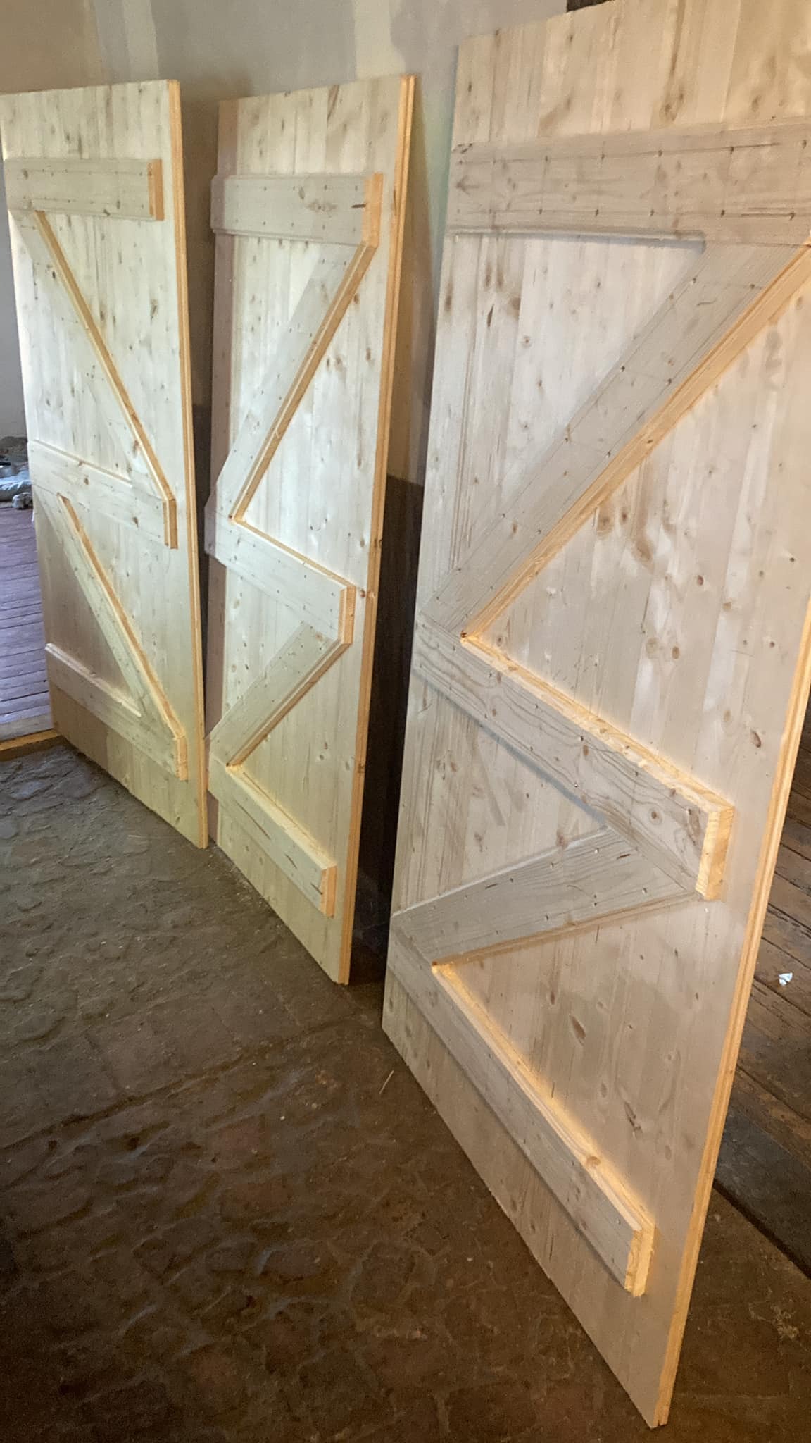 Trzy surowe, drewniane drzwi przesuwne w stylu stodoły, z wyraźnymi słojami drewna i ukośnymi wzmocnieniami, stojące na betonowej podłodze.