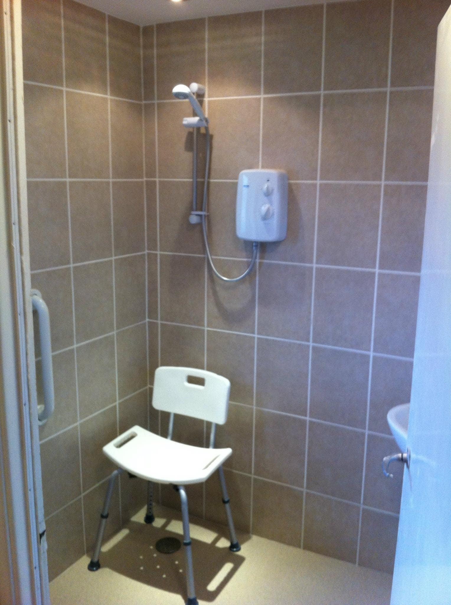 Prysznic z siedziskiem dla osoby niepełnosprawnej, beżowe płytki, biały podgrzewacz wody, uchwyt przy wejściu.