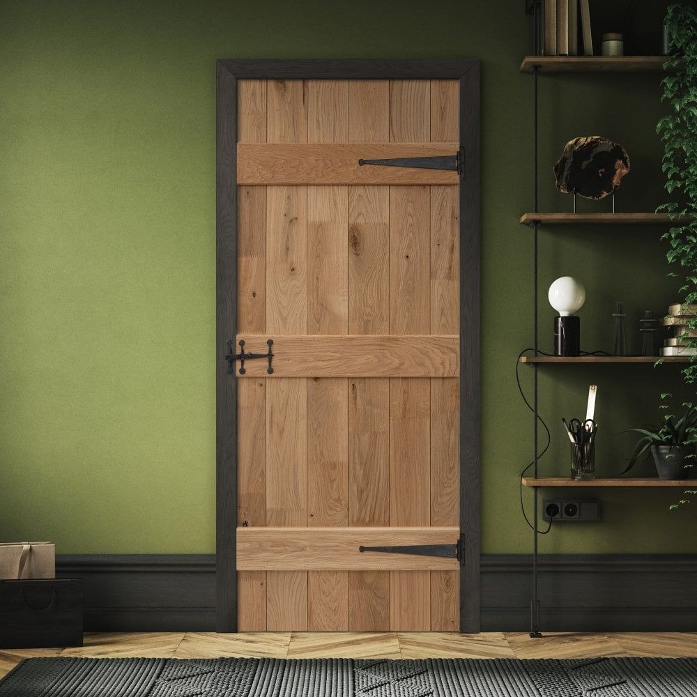 Solidna drewniana drzwi z czarnymi okuciami w rustykalnym stylu, na tle zielonej ściany i drewnianej podłogi, z półkami z dekoracjami w tle.