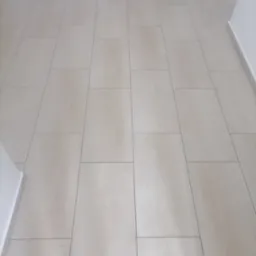 Jasne, podłużne płytki podłogowe ułożone wzdłuż korytarza, imitujące drewno, z widocznymi fugami i białymi listwami przypodłogowymi.