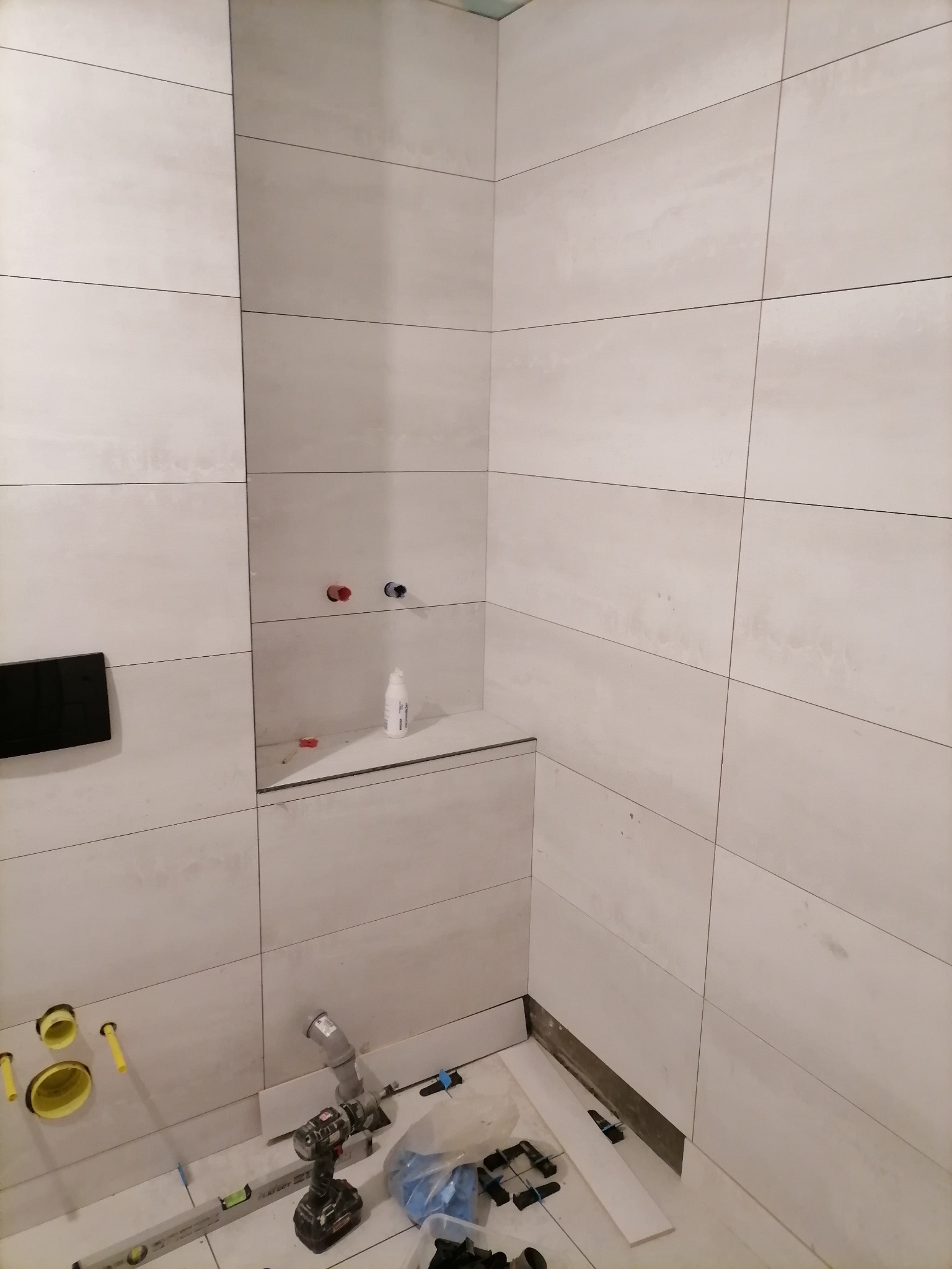 Łazienka w trakcie wykończenia: ułożone jasne płytki imitujące kamień, widoczne przyłącza wodne, stelaż podtynkowy WC, poziomica, wiertarka i elementy instalacji kanalizacyjnej w dolnej części kadru.