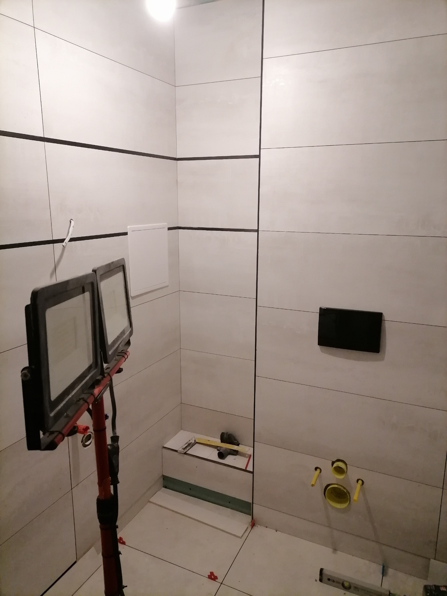 Łazienka w trakcie wykańczania, poziome płytki w odcieniach bieli z ciemnymi fugami, widoczne punkty instalacji sanitarnej, oświetlenie robocze i narzędzia na podłodze.