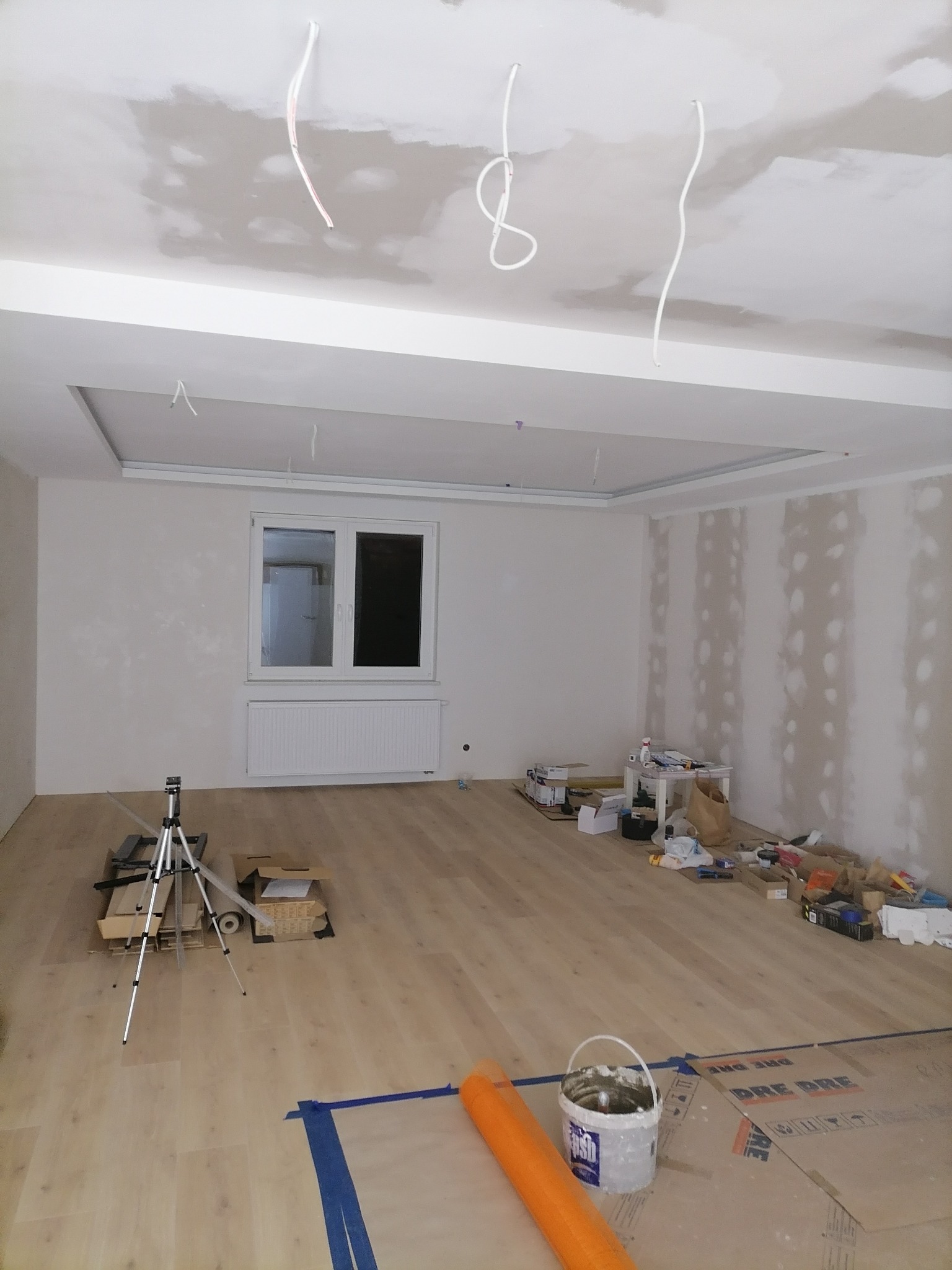 Pokój w trakcie prac wykończeniowych: ściany częściowo pokryte gładzią, widoczne kable zwisające z sufitu, narzędzia i materiały budowlane na podłodze, w tym wiadro z resztkami zaprawy.