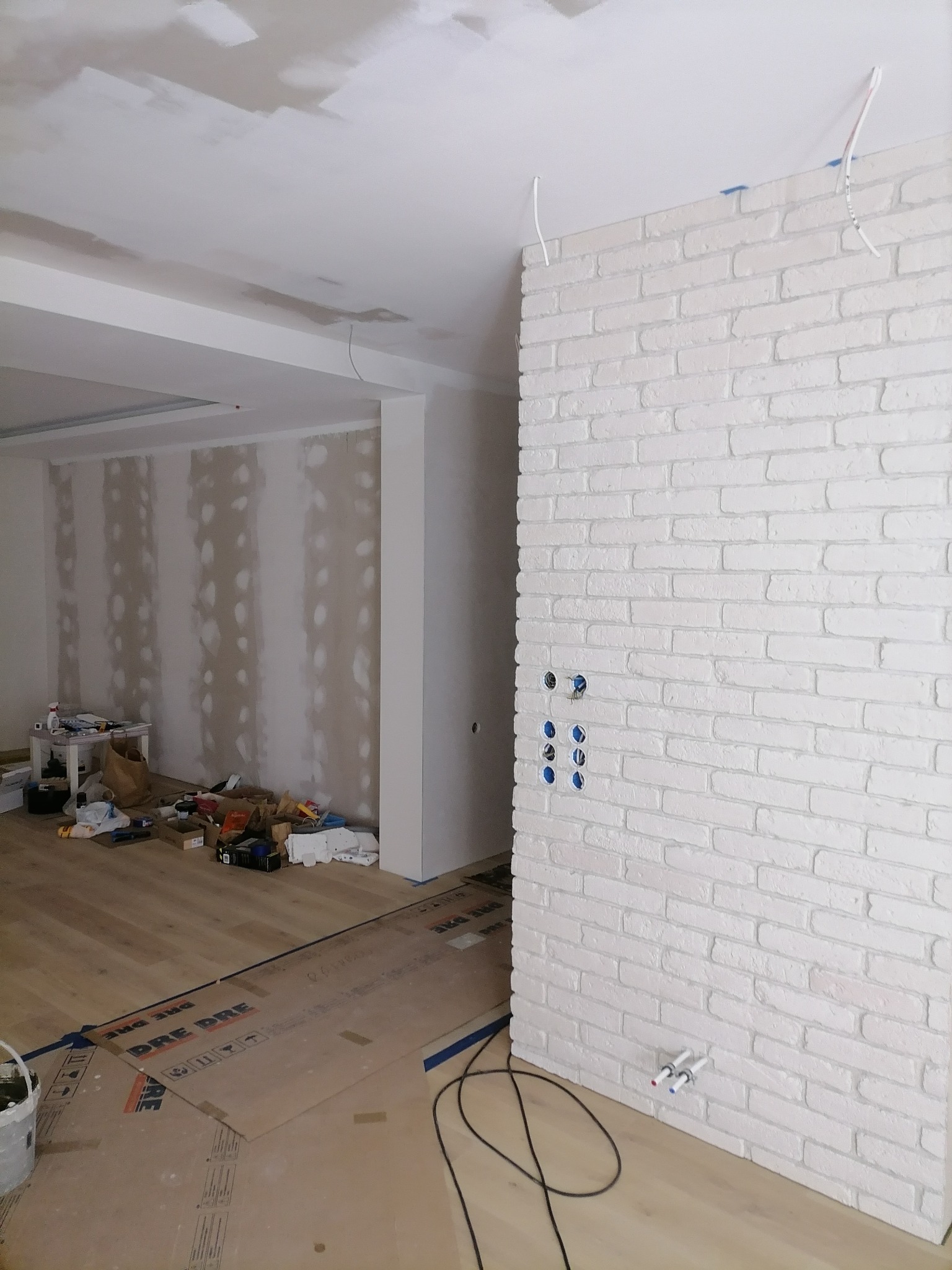 Wnętrze w trakcie wykańczania: ściana z białej cegły dekoracyjnej z przygotowanymi otworami na gniazdka i rury, surowy sufit z widocznymi łatami gładzi, podłoga zabezpieczona kartonem, widoczne...