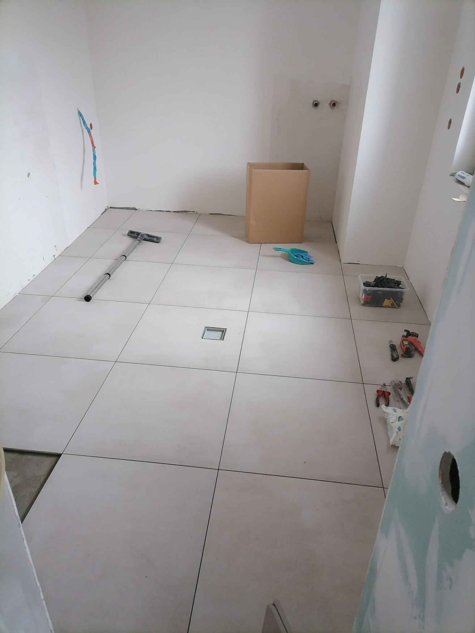 Układanie jasnych, kwadratowych płytek podłogowych w łazience w Strzelcach Opolskich, widoczny odpływ liniowy, narzędzia glazurnicze i kartonowe pudło w tle.