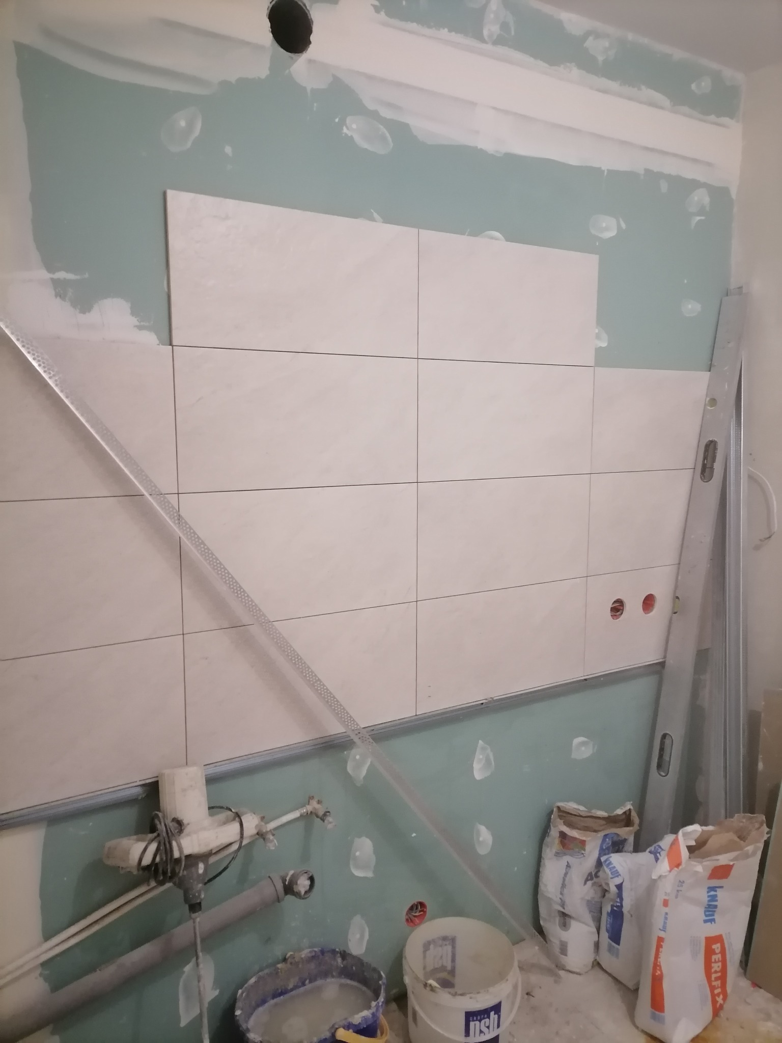 Układanie jasnych, prostokątnych płytek na ścianie z widocznymi puszkami elektrycznymi, przy użyciu poziomicy i profili, z workami zaprawy budowlanej i wiadrem z klejem w tle.