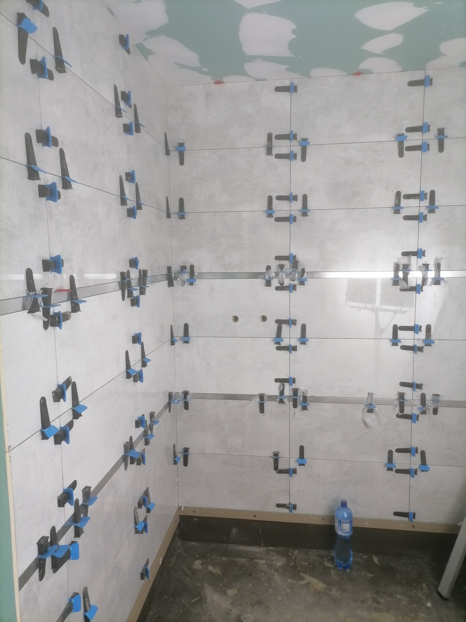 Świeżo ułożone, jasnoszare płytki ścienne w trakcie fugowania, z użyciem systemu poziomowania płytek z niebieskimi klipsami, widoczna butelka wody na podłodze.