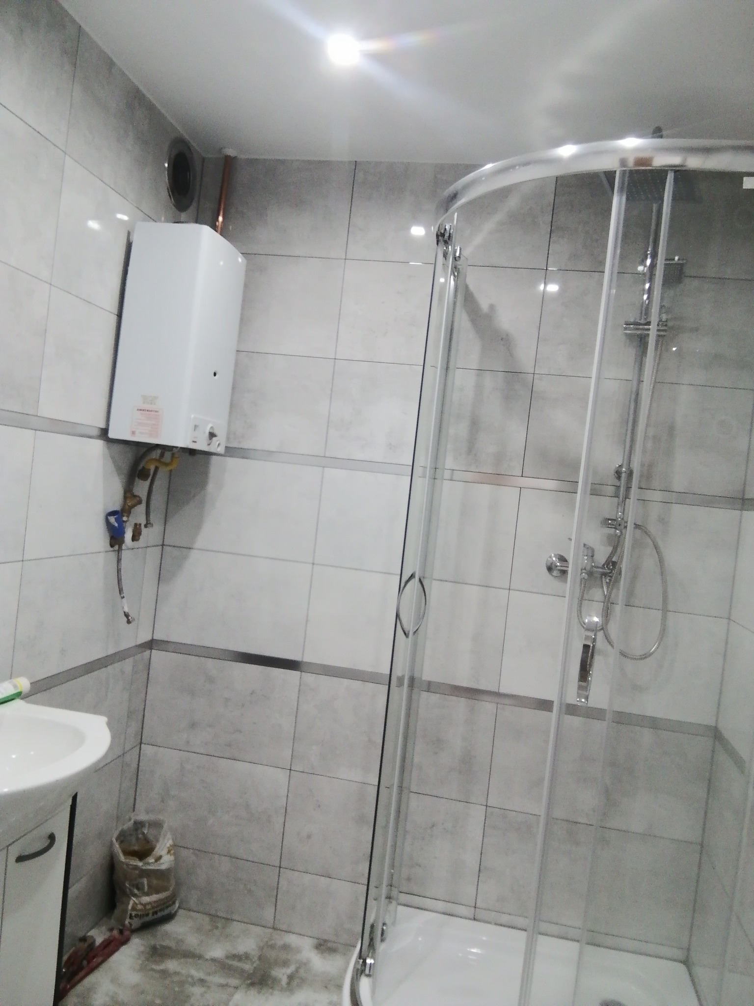 Szara łazienka z kabiną prysznicową, bojlerem na ścianie i workiem z klejem na podłodze.