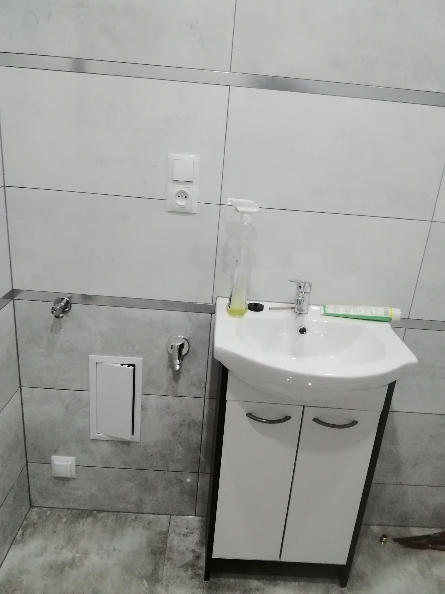 Biała umywalka z szafką w łazience wykończonej szarymi płytkami, widoczne zawory i włącznik światła na ścianie, butelka z rozpylaczem i tuba pasty do zębów na umywalce.