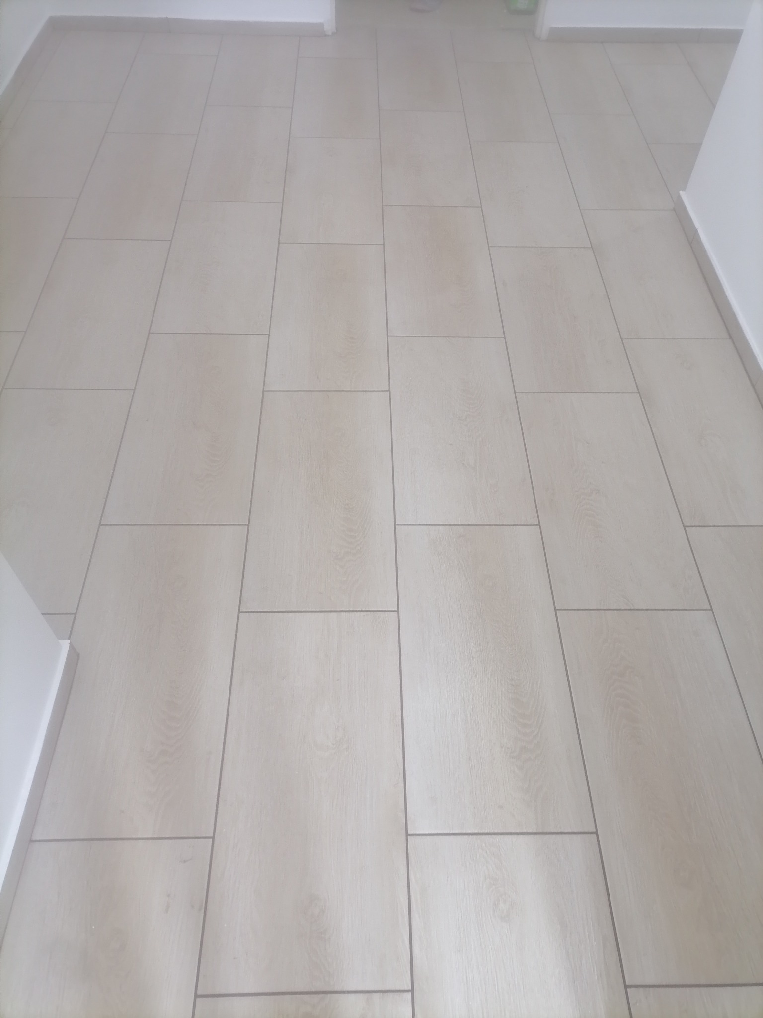 Jasne, podłużne płytki podłogowe ułożone wzdłuż korytarza, imitujące drewno, z widocznymi fugami i białymi listwami przypodłogowymi.