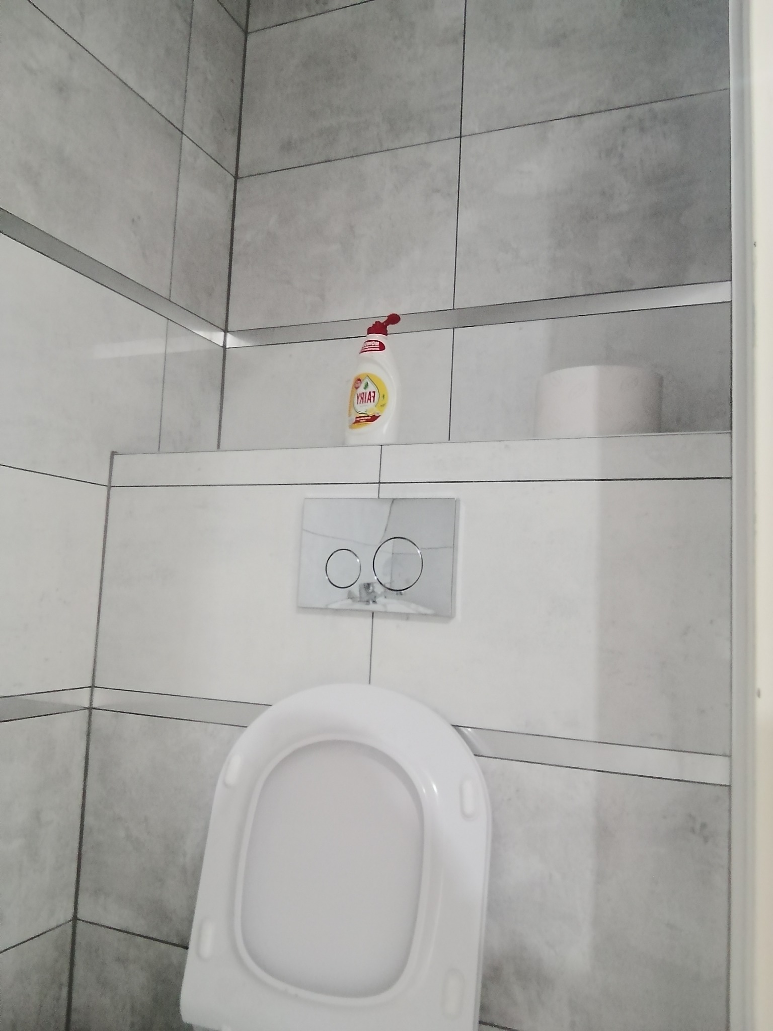 Szare płytki w łazience z wiszącą toaletą i chromowanym przyciskiem spłukującym. Na półce za toaletą stoi płyn do mycia naczyń i rolka papieru toaletowego.